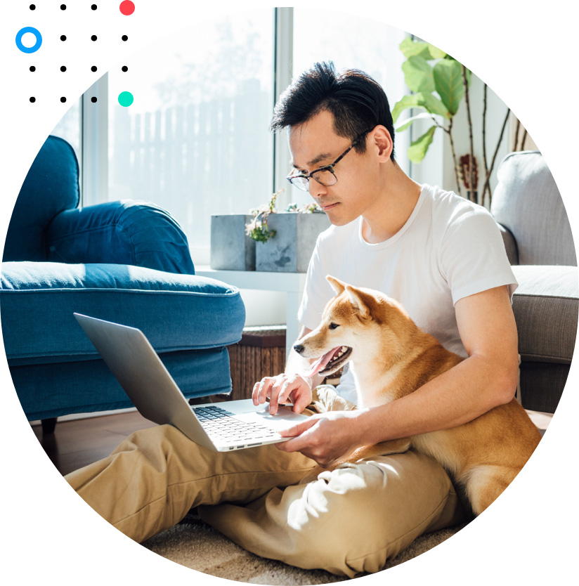 Image of young Asian man, sitting on the floor of his living room by his couch, with his laptop open and dog next to him.