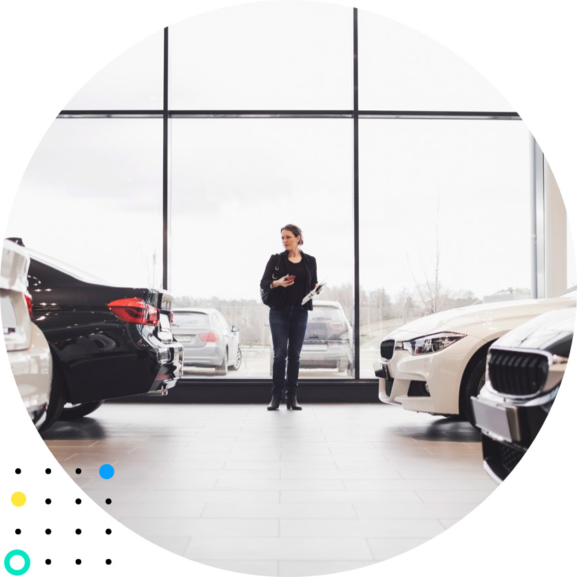 Image of a woman inside a car dealership showroom, phone and printouts in hand, looking at cars.